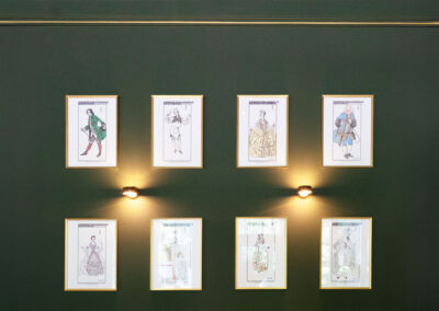 Frisch gestrichene Wand in Lillys Parkcafe mit acht gerahmten Kostümbildern und goldenen Zierleisten.